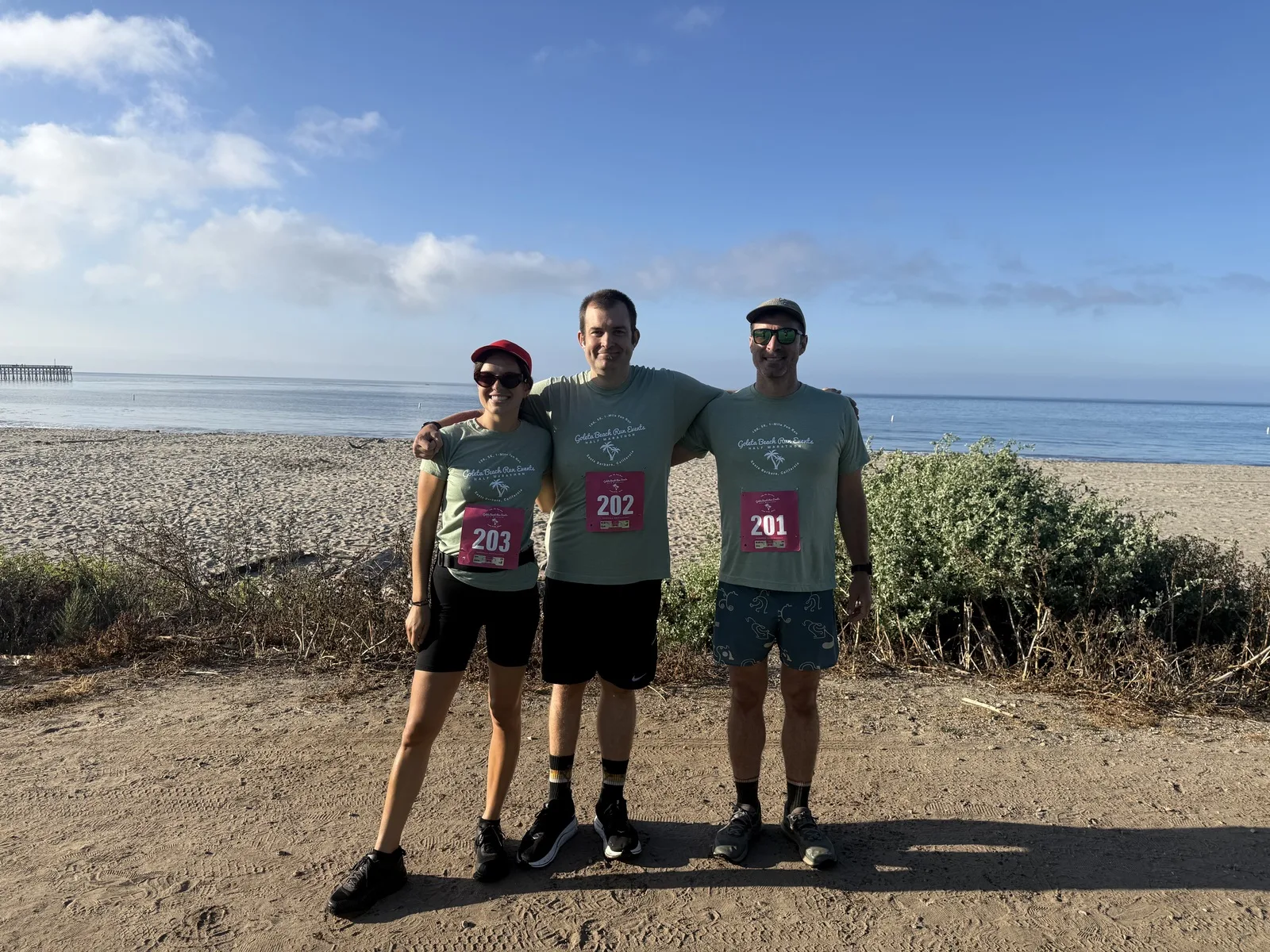 Kevin and two running friends celebrating after a beach 5K, the Pacific Ocean and a pier behind them